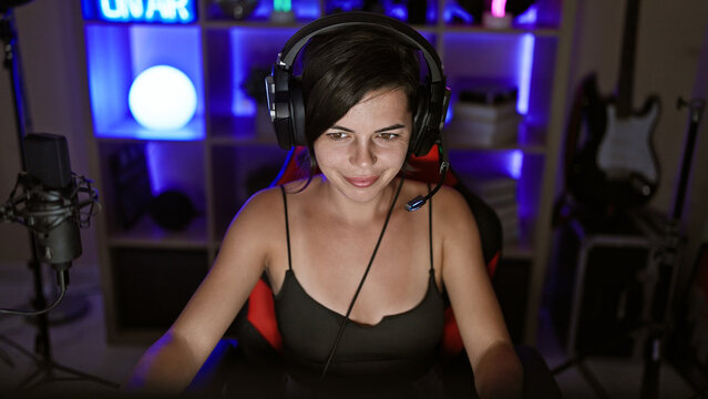 Confident young hispanic woman, beautiful streamer, engrossed in game, streaming in dark gaming room, technology glowing, wearing headphones, ebullient smile lighting up the night