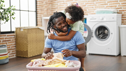 African american father and daughter hugging each other holding clothes of basket sitting on floor at laundry room - obrazy, fototapety, plakaty