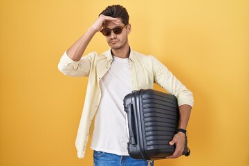 Young hispanic man holding suitcase going on summer vacation worried and stressed about a problem...