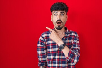 Young hispanic man with beard standing over red background surprised pointing with finger to the side, open mouth amazed expression.