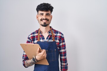 Young hispanic man with beard wearing waiter apron holding clipboard with a happy and cool smile on...