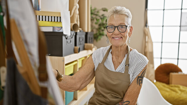 Confident grey-haired senior woman artist, at a fun art studio, smiling as she draws with joy, cultivating her hobby indoors, surrounded by paintbrushes and canvas.