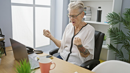 Elegant grey-haired senior woman, a smiling business worker, celebrates a job win at office desk, laptop crossed in a winning gesture.