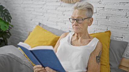 Grey-haired senior woman engrossed in a book, comfortably resting in bed, surrounded by the...
