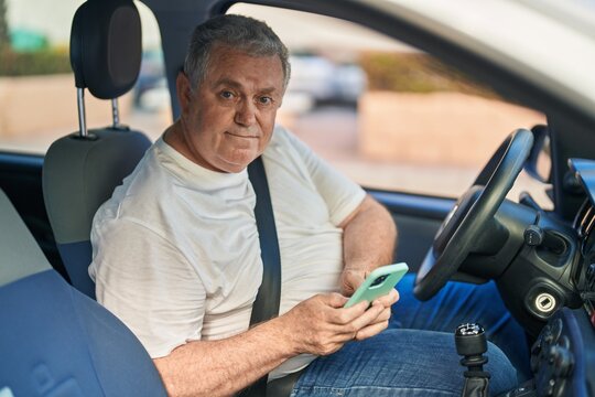 Middle age grey-haired man using smartphone sitting on car at street