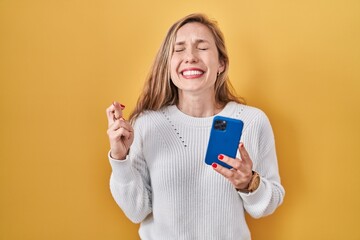 Young blonde woman using smartphone typing message gesturing finger crossed smiling with hope and eyes closed. luck and superstitious concept.