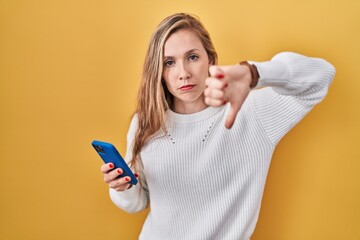 Young blonde woman using smartphone typing message looking unhappy and angry showing rejection and...