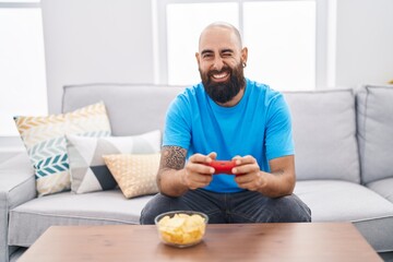 Young hispanic man with beard and tattoos playing video game sitting on the sofa winking looking at...