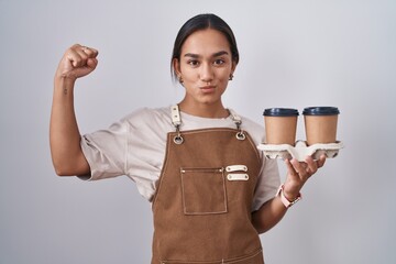 Young hispanic woman wearing professional waitress apron holding coffee strong person showing arm...