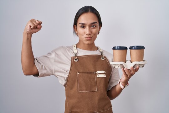 Young hispanic woman wearing professional waitress apron holding coffee strong person showing arm muscle, confident and proud of power