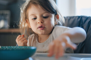 Cute little child with bowl of tasty yogurt in high chair at home.