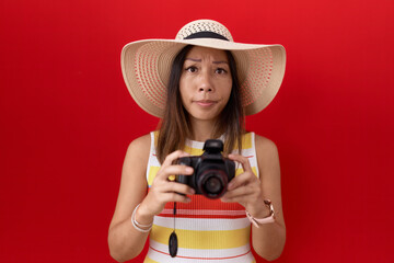 Middle age chinese woman using reflex camera wearing summer hat skeptic and nervous, frowning upset...