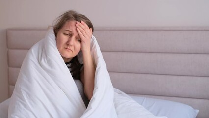 Woman feels bad at home, sitting in bed under white blankets and frowning, touching her head.