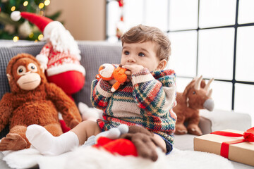 Adorable hispanic toddler sucking doll sitting on sofa by christmas tree at home