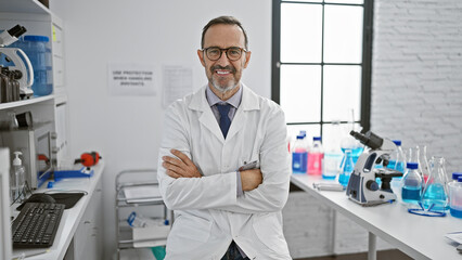 Happy mature, grey-haired male scientist with a confident smile enjoys his research work, sitting at his lab table, arms crossed in a welcoming gesture, connected with science and medicine. - obrazy, fototapety, plakaty