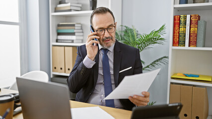 Energetic middle age man with grey hair, immersed in his professional role at the office, confidently talking on a smartphone while attentively reading a crucial business document.