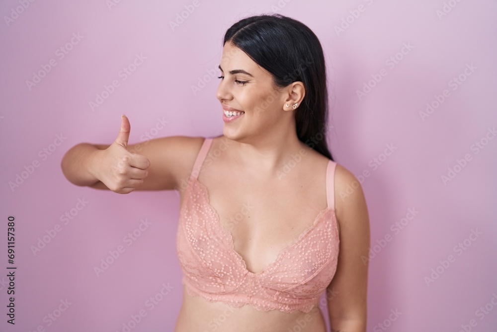 Wall mural Young hispanic woman wearing pink bra looking proud, smiling doing thumbs up gesture to the side