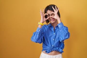 Young girl standing over yellow background doing ok gesture like binoculars sticking tongue out, eyes looking through fingers. crazy expression.