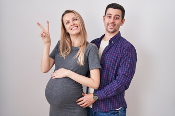Young couple expecting a baby standing over white background smiling with happy face winking at the...