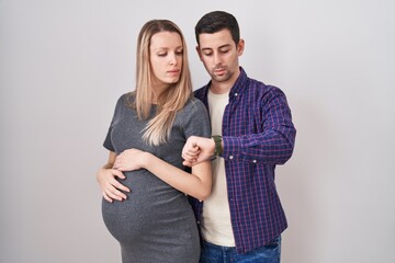 Young couple expecting a baby standing over white background checking the time on wrist watch,...