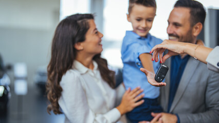 Happy parents with small kid after receiving keys for their new car in a showroom. Congratulations,...