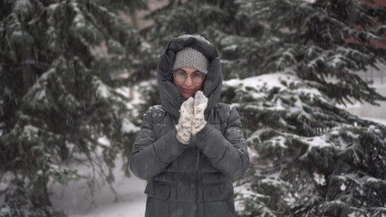 A young woman stands against a background of fir trees under heavy snowfall and rubs her hands...