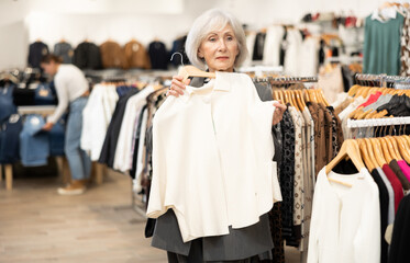 Happy old woman customer choosing white cardigan in clothing shop with large assortment