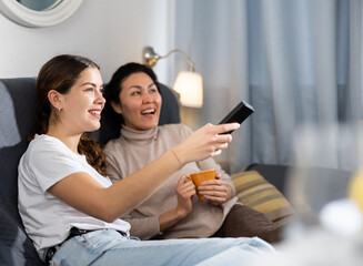 Positive Asian and European women sitting on couch and watching TV together.
