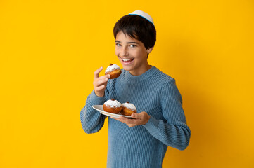 Little boy in kipa holding plate with tasty donuts on yellow background. Hanukkah celebration