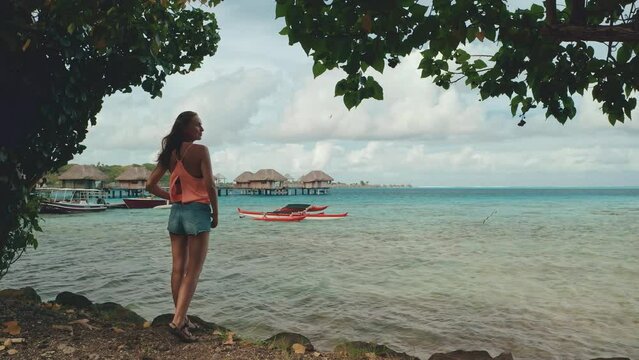 Woman stand on coast looking at sea with wooden huts and fishing boats on Bora Bora island. Back view