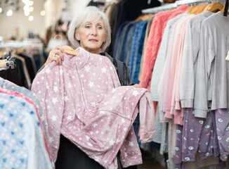 Elderly woman buyer chooses pajamas in clothing store..