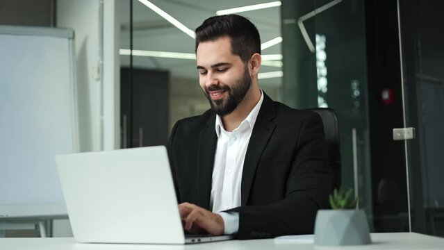 Satisfied bearded man with dark hair and beard wearing stylish black suit happily closing wireless laptop after finishing work at modern office. Concept of people, successful career and technology.