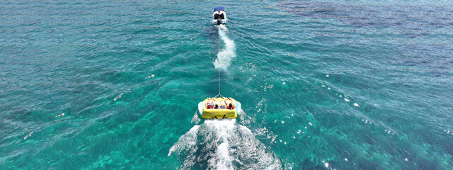 Aerial drone ultra wide panoramic photo with copy space of children riding inflatable tow round donuts pulled by powerboat in high speed known as water sports in deep blue sea