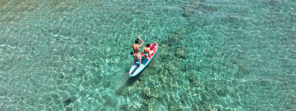 Aerial drone ultra wide photo of couple sitting on SUP or Stand Up Paddle board cruising in low speed in tropical paradise exotic bay with emerald crystal clear calm sea
