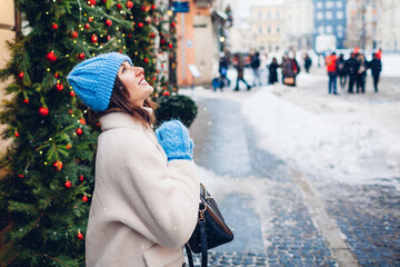 Happy young woman admires falling snow walking on European city street decorated for Christmas...