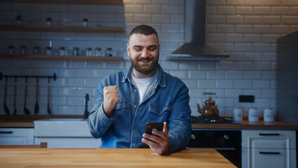 Bearded young man sitting against the kitchen counter checking messages read notice receive good...