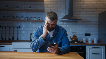 Bearded young adult man sitting against the kitchen counter having headache while using smartphone. Tired young man get having pain from a headache or migraine	 - obrazy, fototapety, plakaty