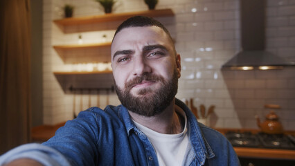Fototapeta premium Selfie of cozy young bearded adult man holding a smartphone in hand looking at camera making video or selfie in kitchen. Modern tech, self leisure, enjoying, having fun