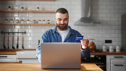 Bearded young adult man sitting against the kitchen counter entering credit card number on laptop...