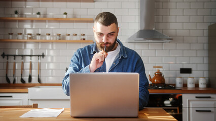 Bearded young adult man typing on laptop doing research sitting against the kitchen working on...