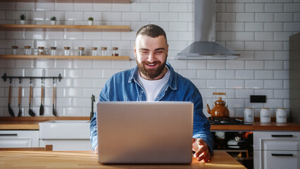 Smiling young adult bearded man sitting against the kitchen counter having video call conversation through using laptop. Share news, talk to family, enjoy webchat virtual meeting	