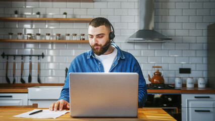 Bearded young adult business man wear wireless headset sitting against the kitchen counter having...