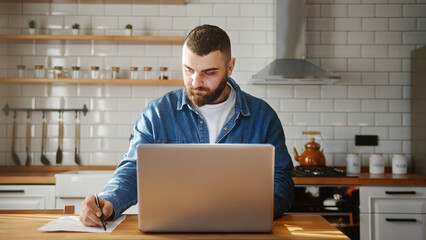 Bearded young adult man wearing in-ear headphones typing on laptop doing research sitting against...