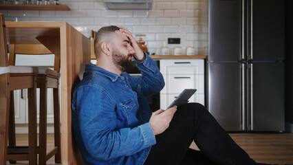 Young man sitting on the floor leaning against the kitchen using tablet device having anxiety and...