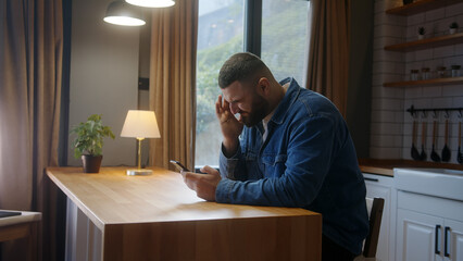 Bearded young man sitting against the kitchen counter having headache while using smartphone. Tired...