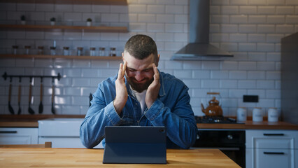 Bearded young adult man sitting against the kitchen counter having headache while using tablet...
