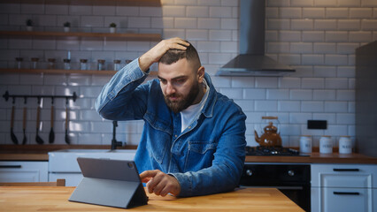 Bearded young man sitting against the kitchen counter using tablet device having anxiety and stress. Upset man reacting to loss, bad news - obrazy, fototapety, plakaty