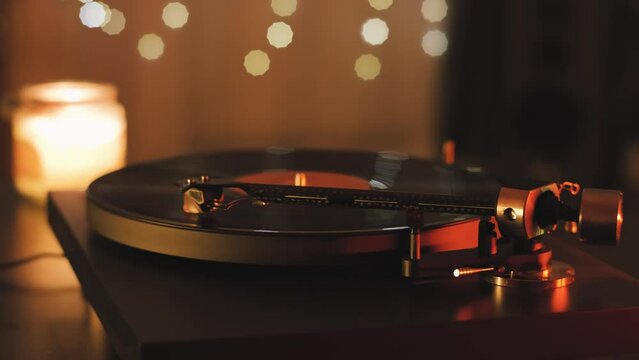 A vinyl record spins in the modern gramophone music player and plays an old disco. Close-up shot of custom vinyl turntable player with carbon tonearm. Candle and garlands in the background