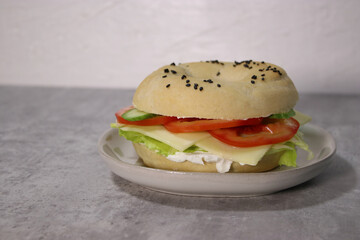 Delicious bagel with vegan cheese and fresh vegetables on a plate, grey table blurry background