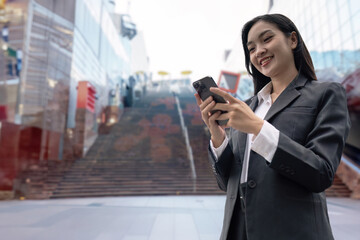 Asian businesswoman using mobile phone while walking to office, skyscrapers cityscape background